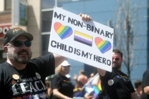 A white man with a beard in a "Pride" hat and sunglasses holds up a hand-written sign reading, "My non-binary child is my hero." The sign includes two rainbow-striped hearts and a non-binary pride flag with yellow, white, purple and black stripes.